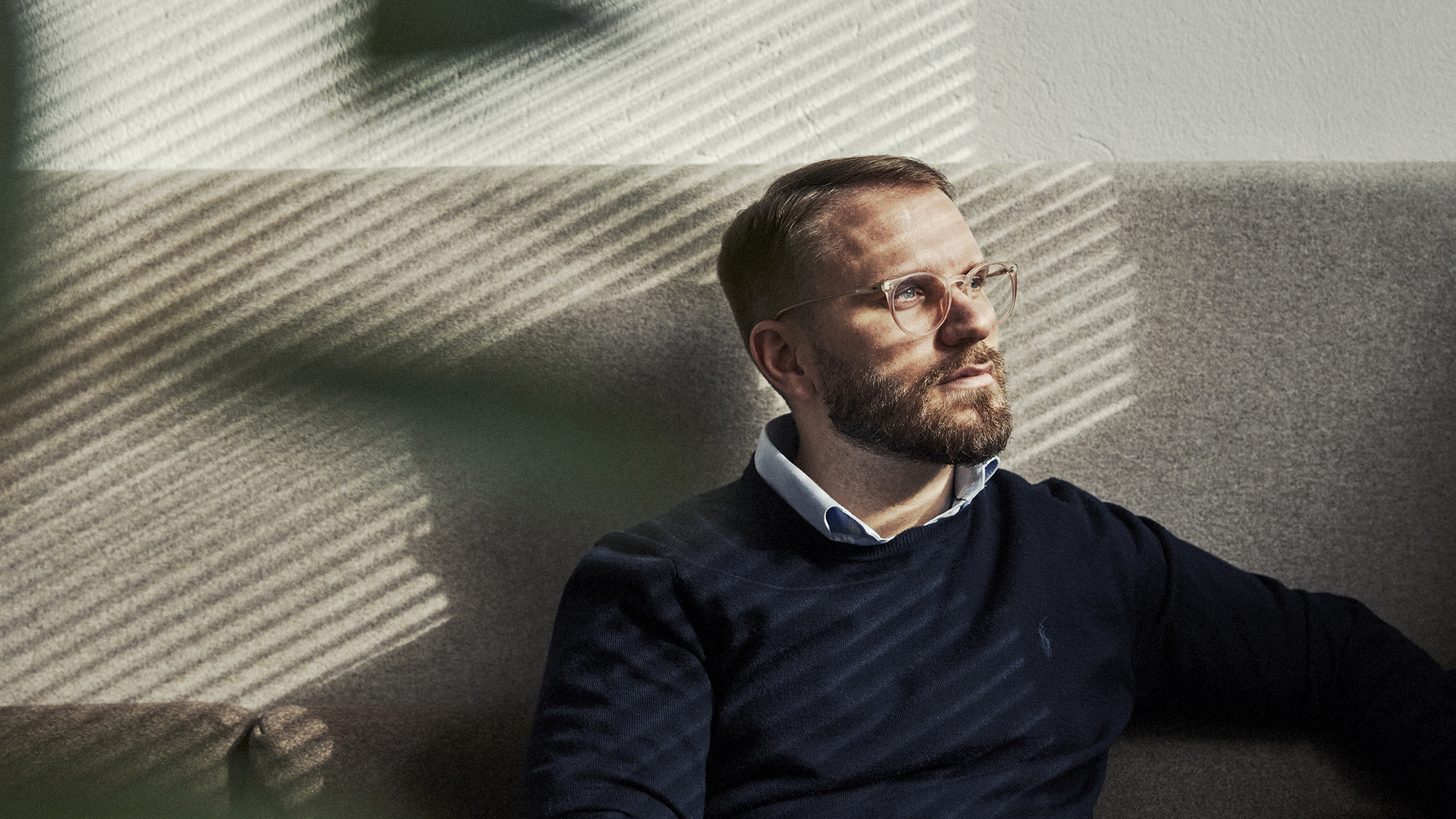 En man med skägg och glasögon sitter lutad mot en vägg, belyst av strimmor av ljus från persienner.